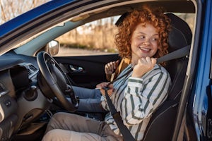 A teenage girl sitting in the driver's seat of a blue car. She's looking behind her, smiling, and fastening her seat belt.