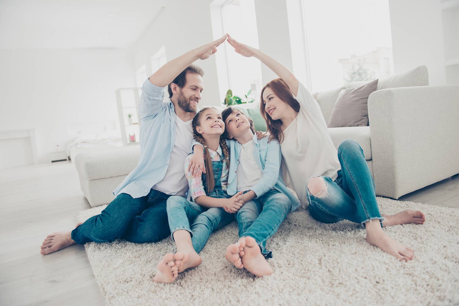 Two children sit on the carpet while their parents form a roof shape with their arms over their heads.