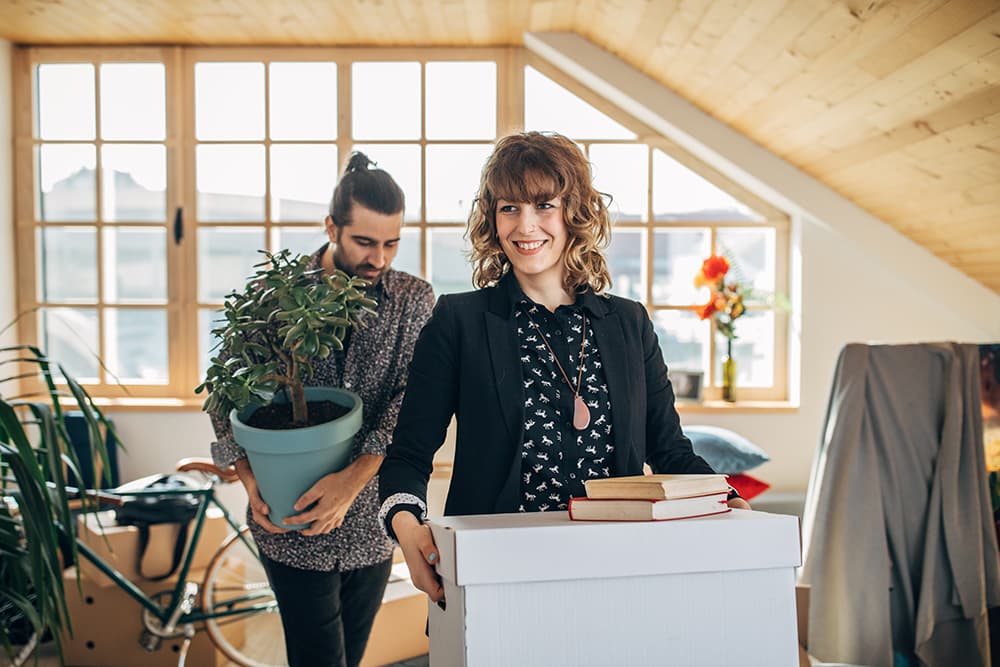 A cheerful woman stands in an empty apartment, holding a cardboard box. Behind her, another person holds a potted plant.