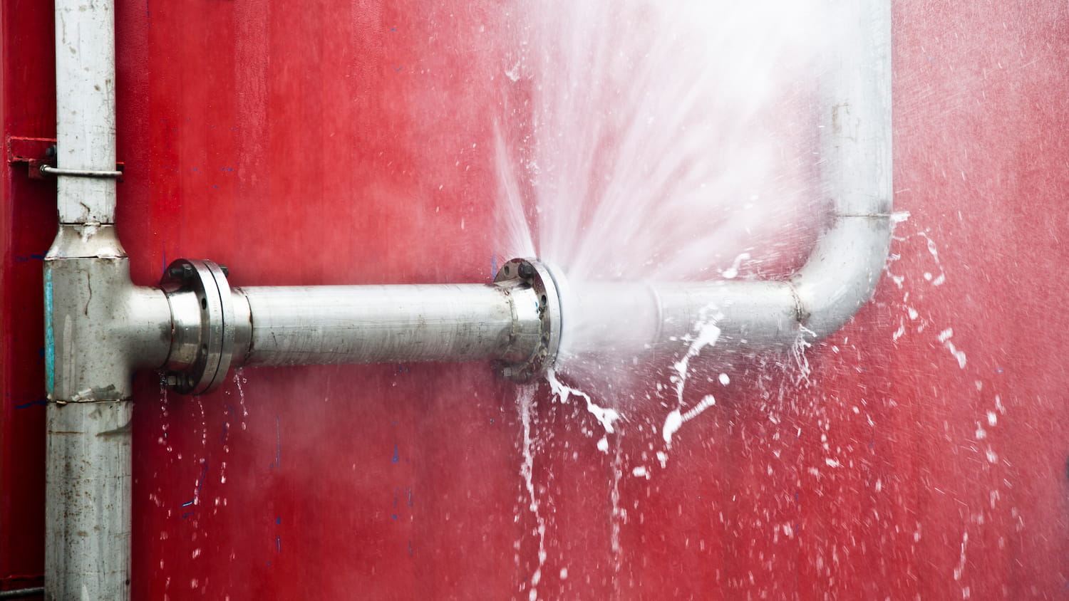 Water spraying from a burst pipe against a red wall.