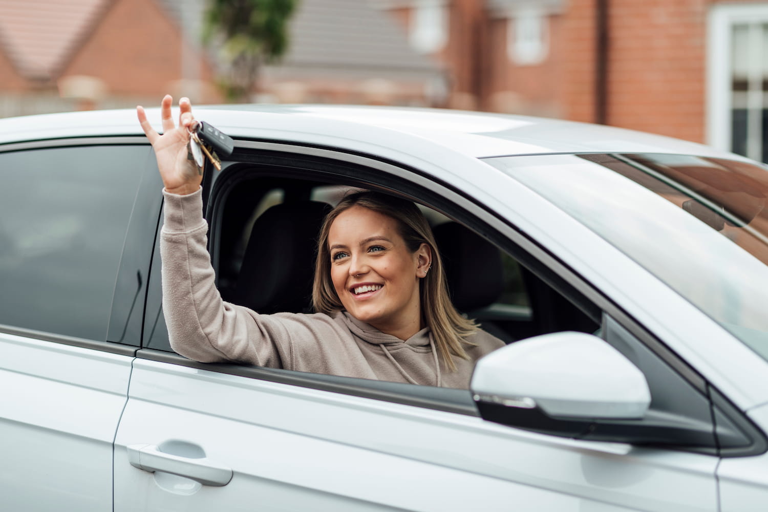 A female leaning out of a car window holding a set of car keys and looking happy as she has passed her driving test.