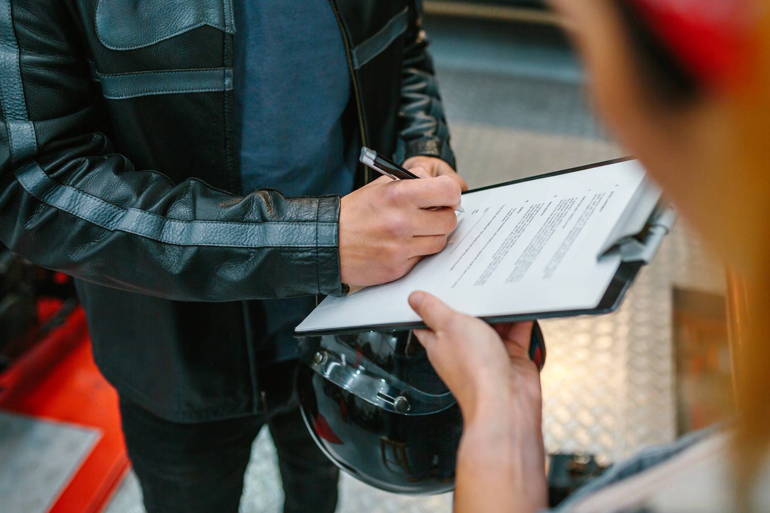 Close-up of a person in a leather jacket signing paperwork for a repaired motorcycle at a workshop, with a staff member holding a clipboard nearby.