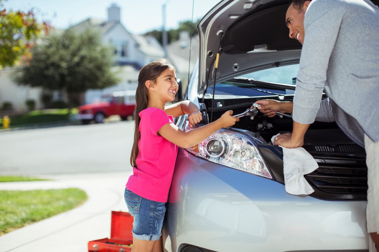 Father and daughter standing in front of a car's open hood. The father is working on the car with one hand and handing the daughter a wrench with the other.