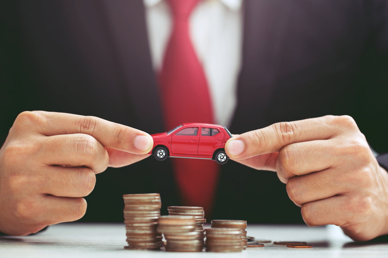 Person in suit and red necktie holding model of toy car on over a lot of stacked coins