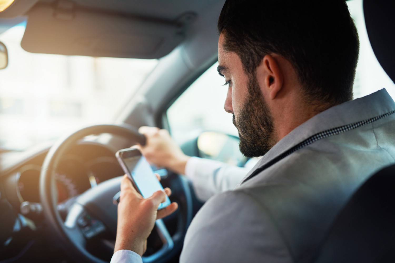 Man sitting in car, looking at phone with one hand on the wheel.