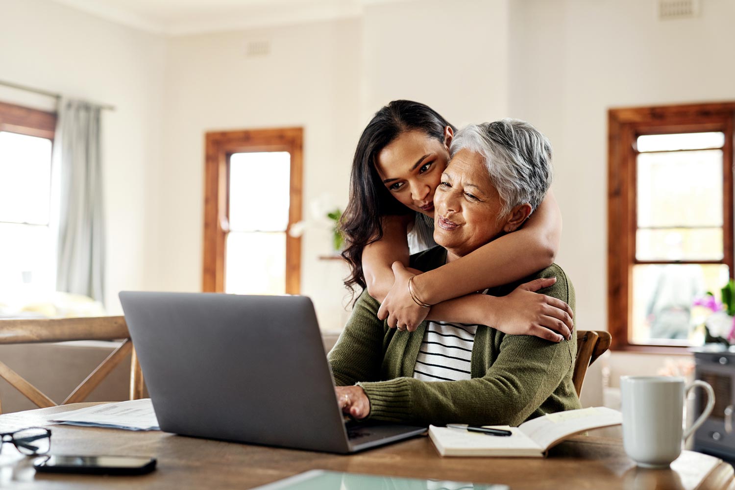 A young individual hugs an older person who is using a laptop.