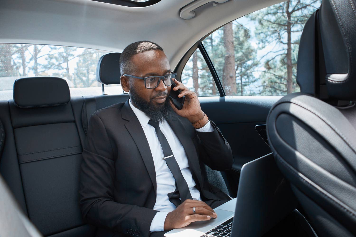 Person in a suit is working on his laptop while sitting in the backseat of a car, talking on his phone.
