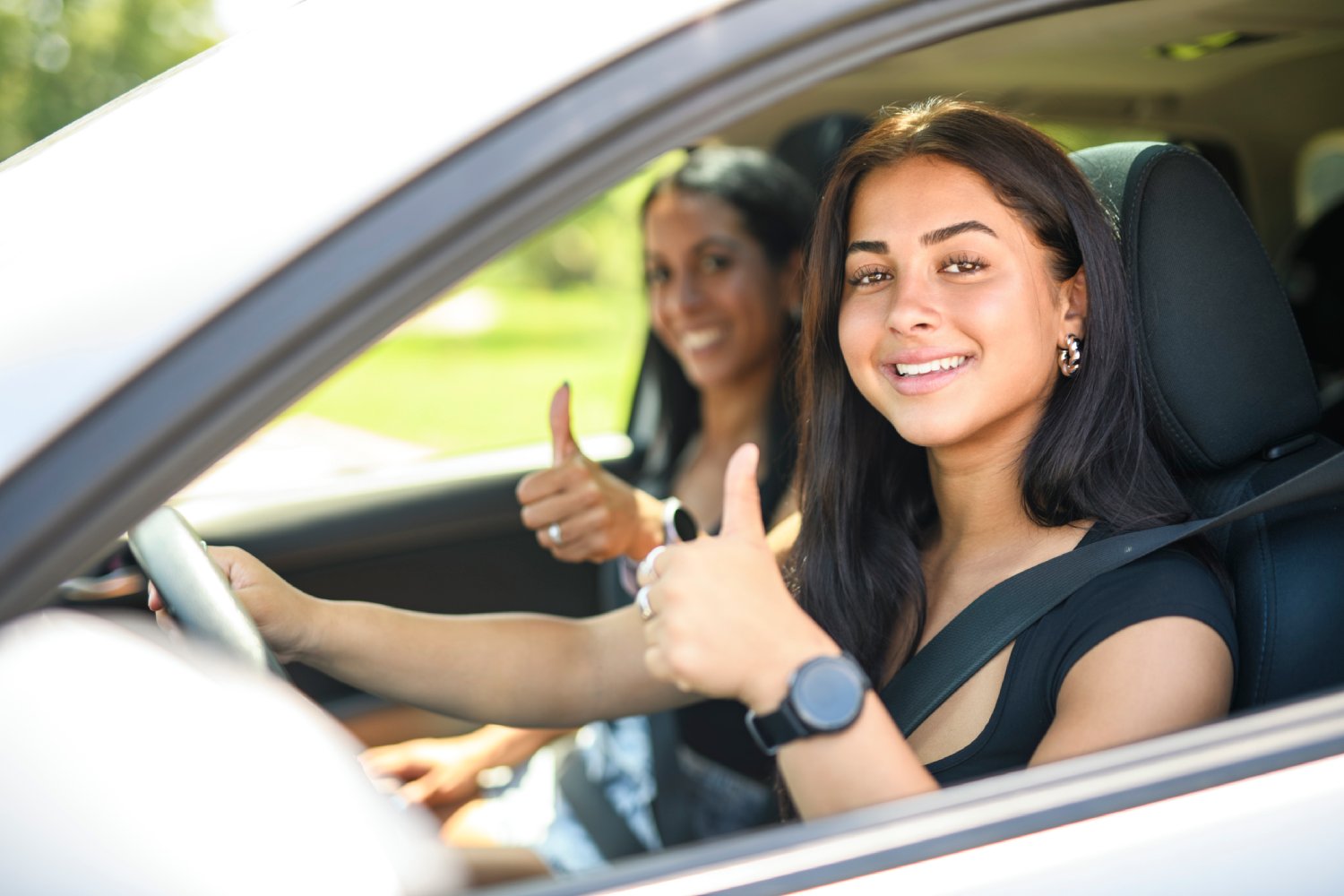Two people sitting in car, smiling and holding a thumbs up.