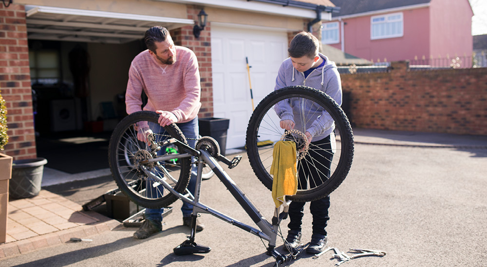 A parent and child collaborate on a bicycle in the driveway or garage.