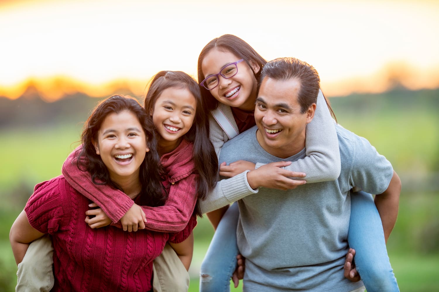 A smiling family of four enjoys piggyback rides outdoors at sunset.