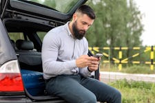 Man sitting in open trunk of his car checking his phone.