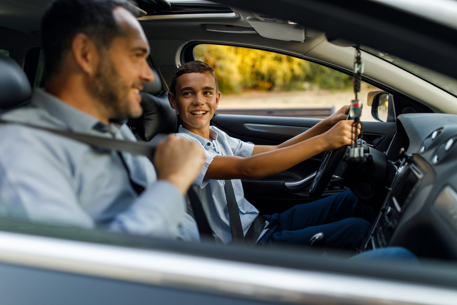 Focus shot of cheerful mid adult man sitting in passenger’s seat and putting on seatbelt next to a teenage son in the driving seat.