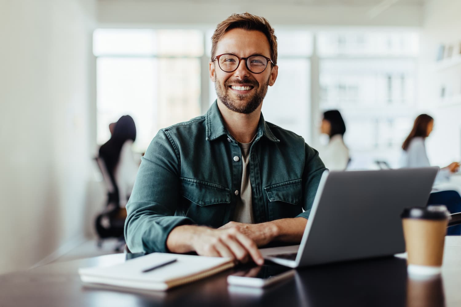 Portrait of a smiling man sitting in an office and working on a laptop with his colleagues in the background.