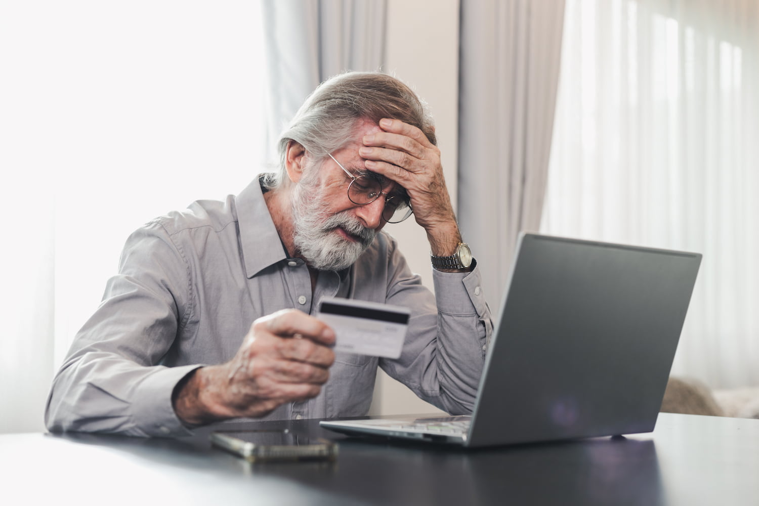 Elderly man at laptop with credit card in hand, frustrated.