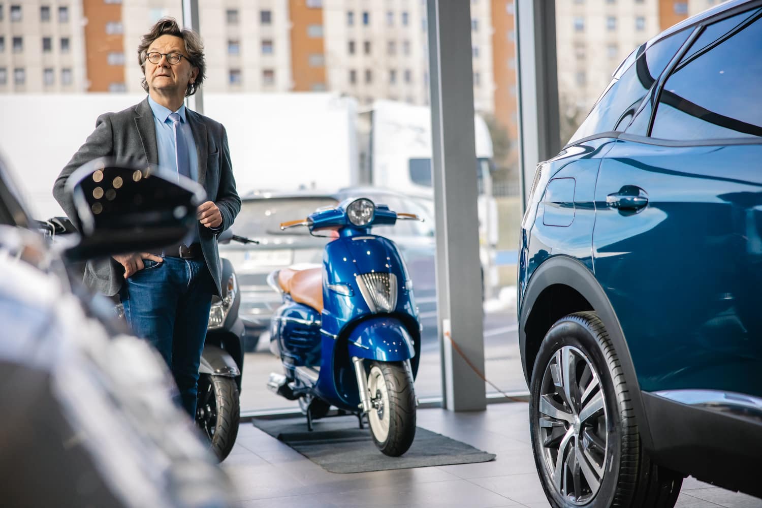 A person in a suit stands beside a new car and bike, looking seriously at a distant person.