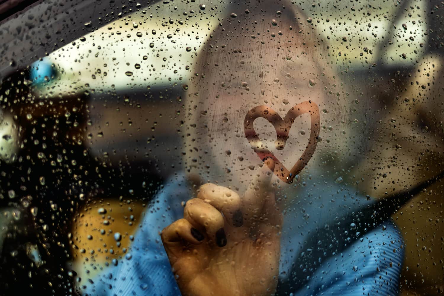 Happy woman drawing a heart on a foggy car window during a rainy day.