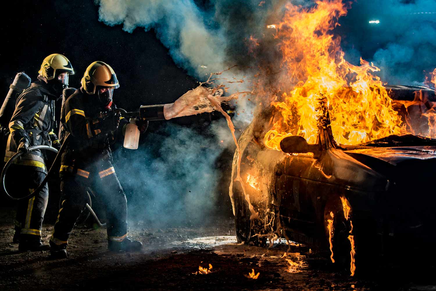 Two firefighters extinguishing a burning car with a foam extinguisher at night.