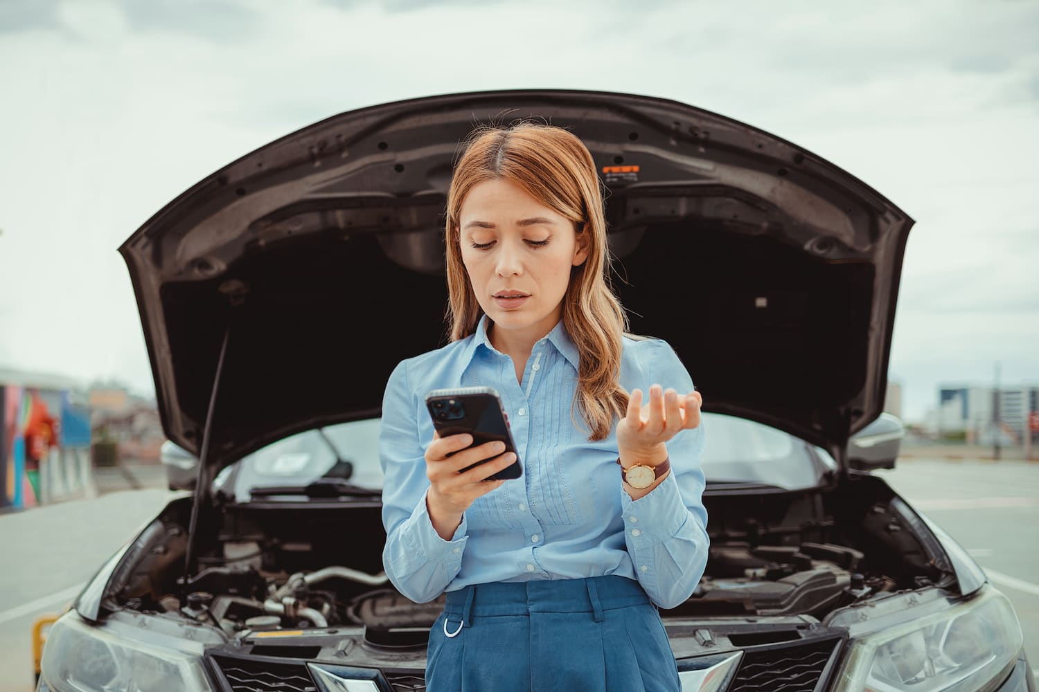 Woman calling autoservice on the phone with an open-hood car behind her