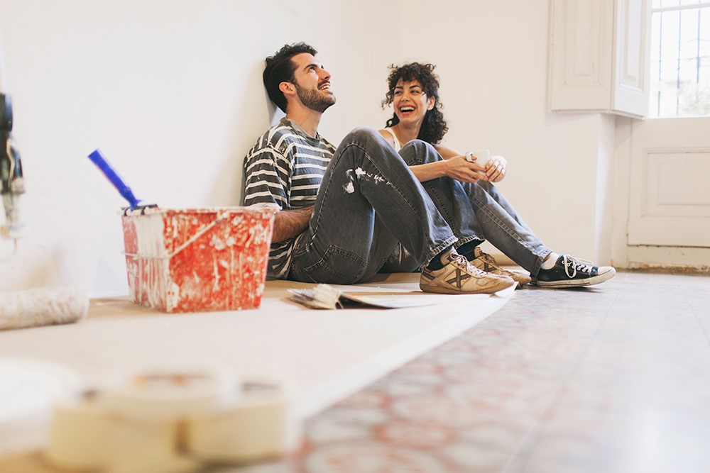 A happy couple sitting on the floor with a paint bucket and brush nearby.