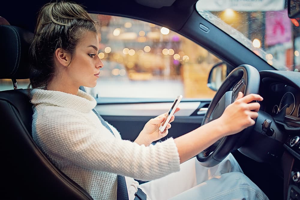 A woman in the driver's seat, looking at her phone with one hand on the steering wheel.
