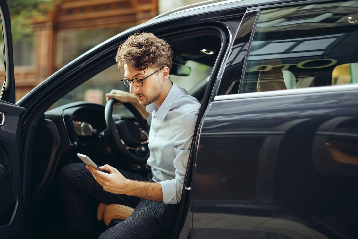 A person sits in the passenger seat of a vehicle with the door open, one leg extended outside, looking at their phone.