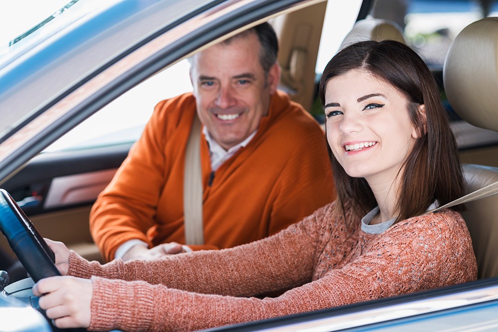 A young woman seated in the passenger seat, smiling and holding the steering wheel, next to an older man who is also smiling.