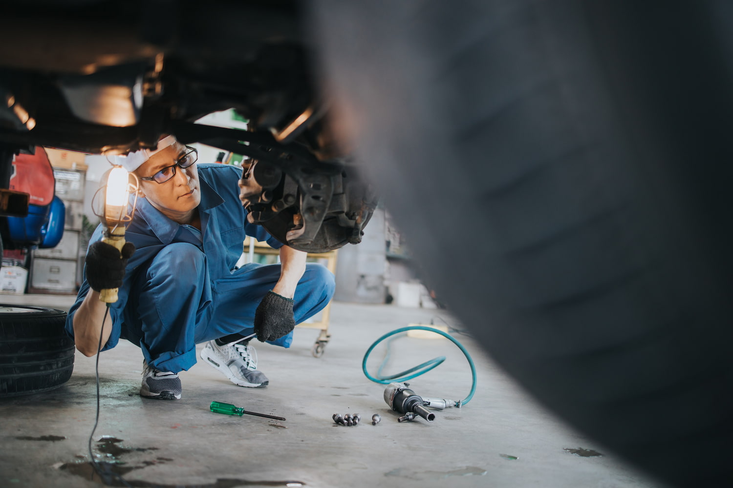 A mechanic in blue coveralls inspects the underside of a vehicle using a handheld work light, surrounded by tools and car parts in a garage.