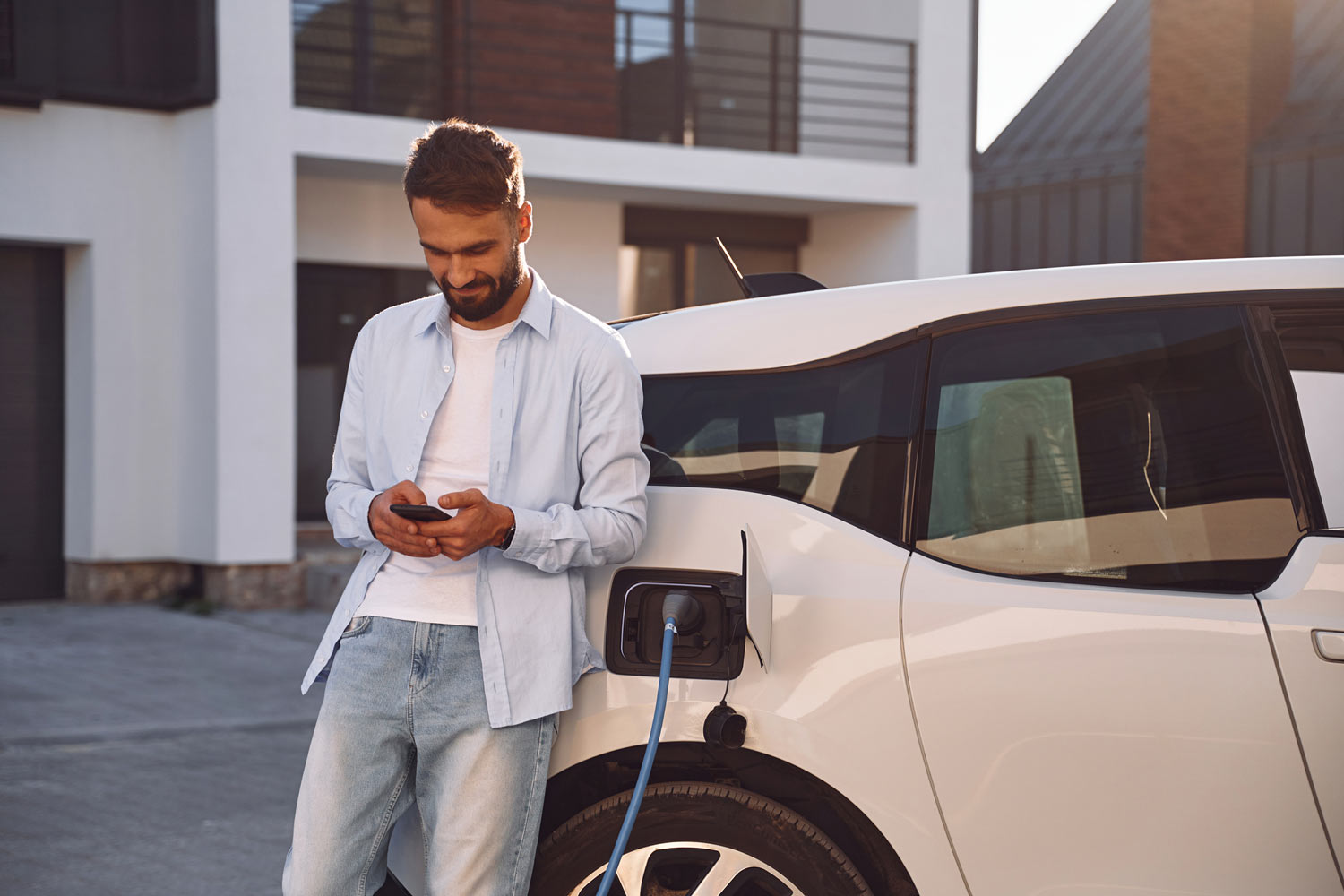 Person charging a car near a house while holding a smartphone.