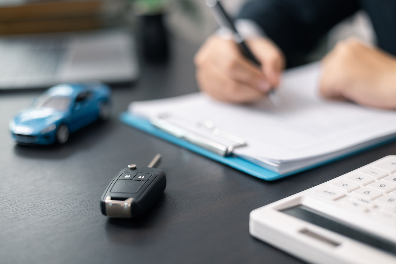 Someone is signing documents with a pen, while a model car and a set of keys rest on the table nearby.