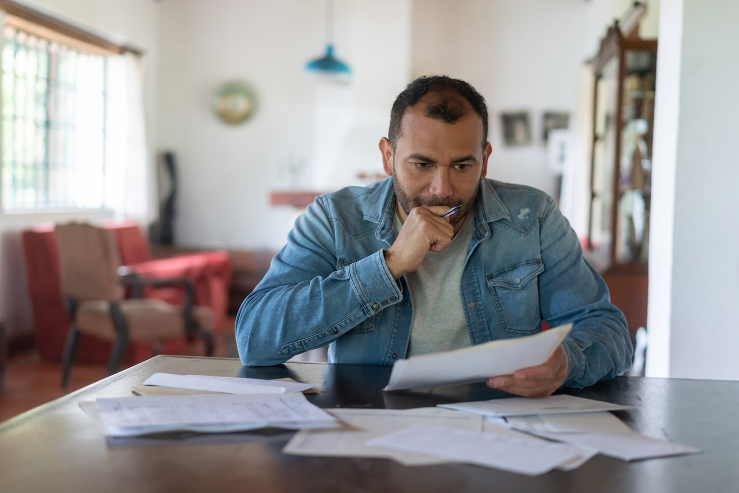 A person sitting at a table with papers scattered around, appearing stressed while holding a sheet of paper.