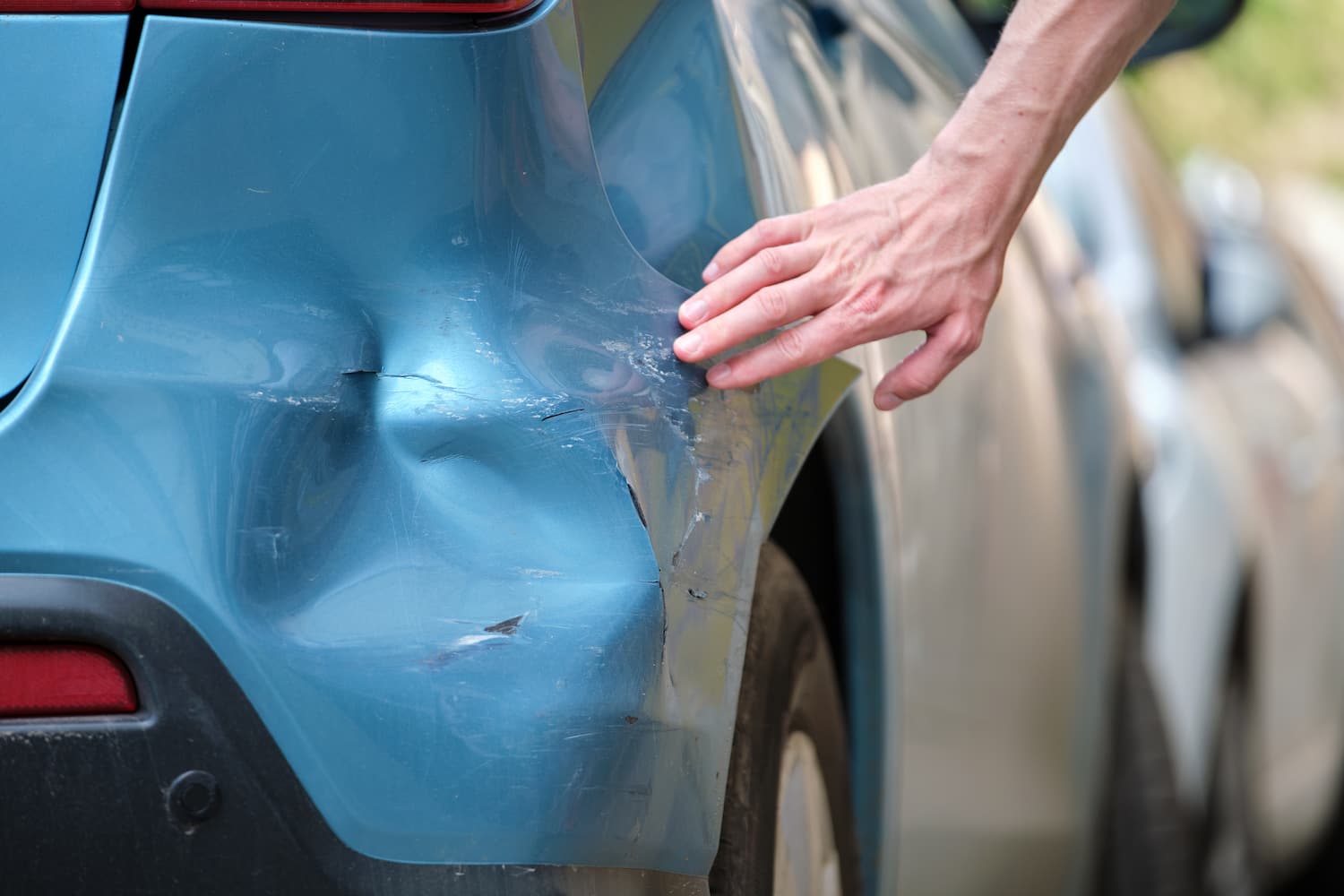 Driver hand examining dented blue car with damaged fender parked on city street side.