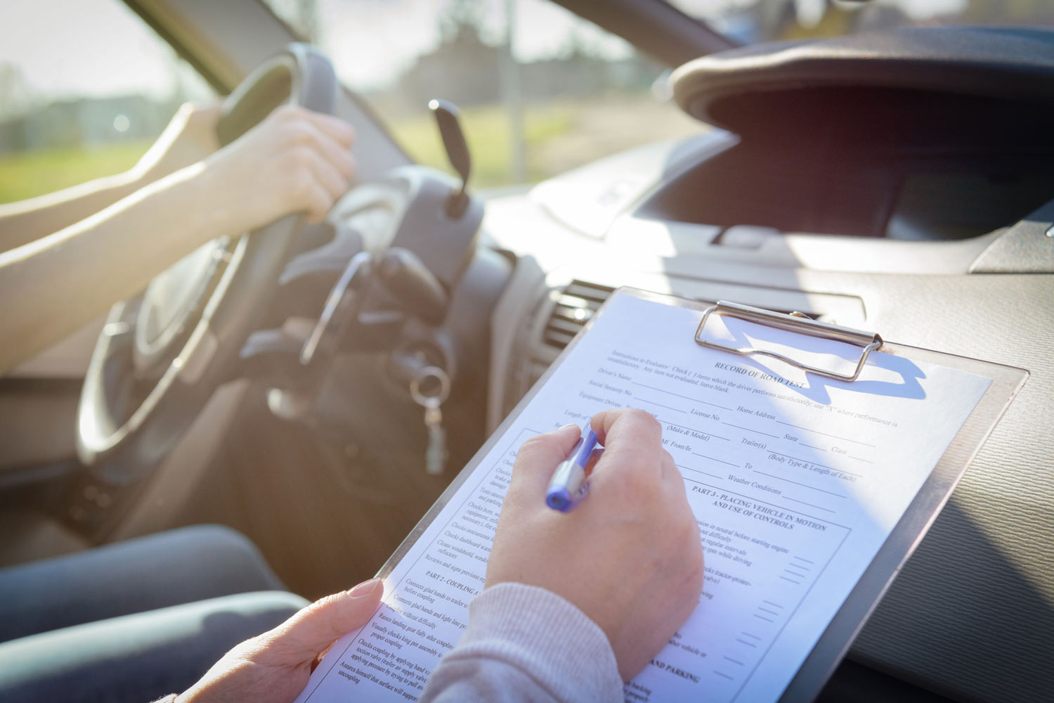 Person filling out a driver's test form while seated in a car with a student.
