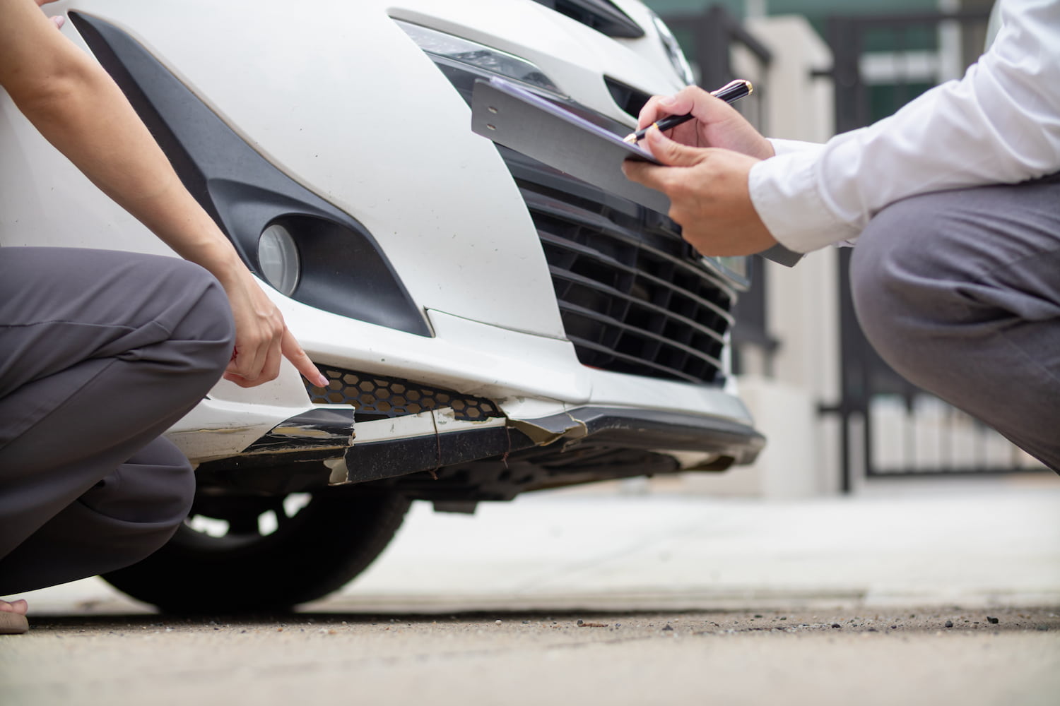 Two individuals are kneeling beside a car, with one person holding a clipboard and pen.