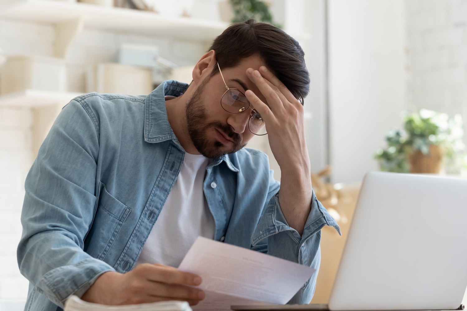 Young man reading a letter paper document sitting at home table and looking depressed