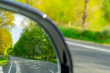 Close-up of a rear-view mirror showing a road surrounded by green and yellow trees