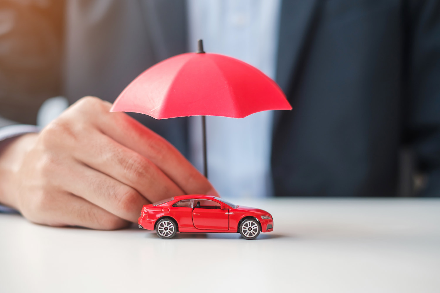 A person's hand holding an umbrella to protect a small red car model on a table.